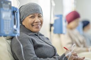 Photograph of a pacific women smiling at the camera. She is sitting hospital room with other patients (blurred in the background) who seem to be receving medicines via a drip. 