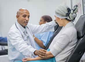 Photograph of a male doctor sitting with a female patient in a ward, with another patient in the background